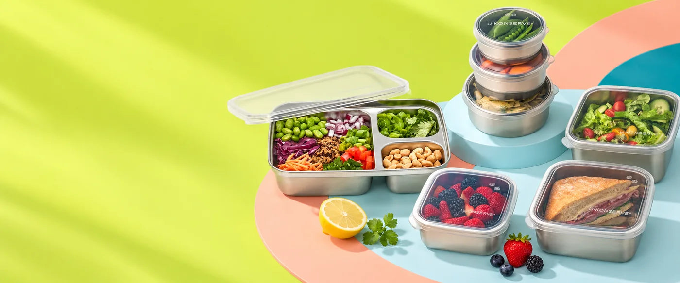 U-Konserve stainless steel containers with clear silicone lids, packed with salad, berries, a sandwich and snacks, on a lime and coral table