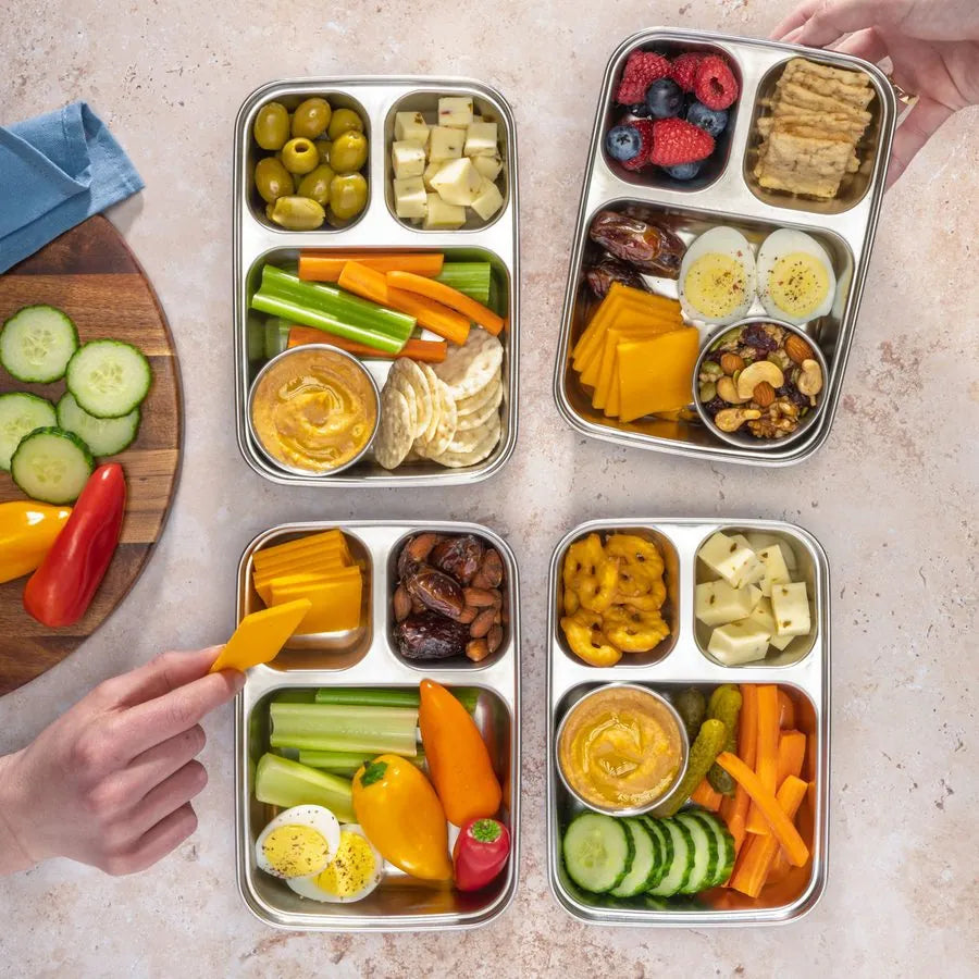 Four divided stainless steel lunch containers prepped with vegetables, fruit, cheese and dips