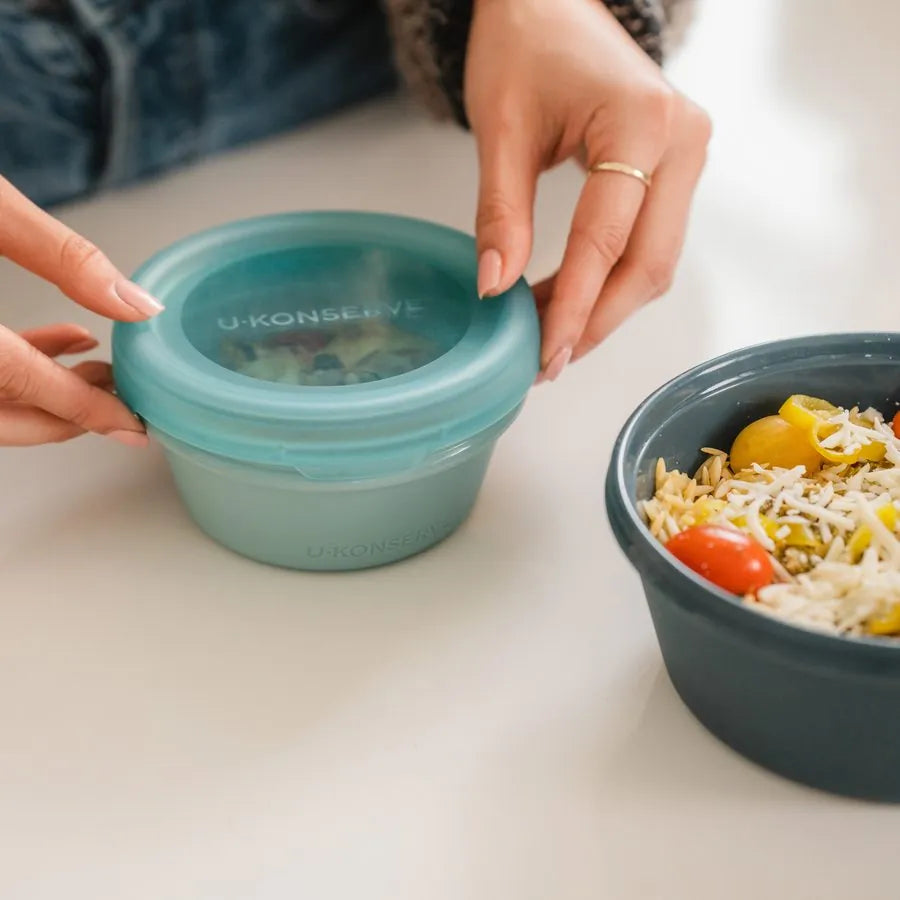 Hands closing a seafoam Bounce Box silicone container next to a bowl of orzo salad