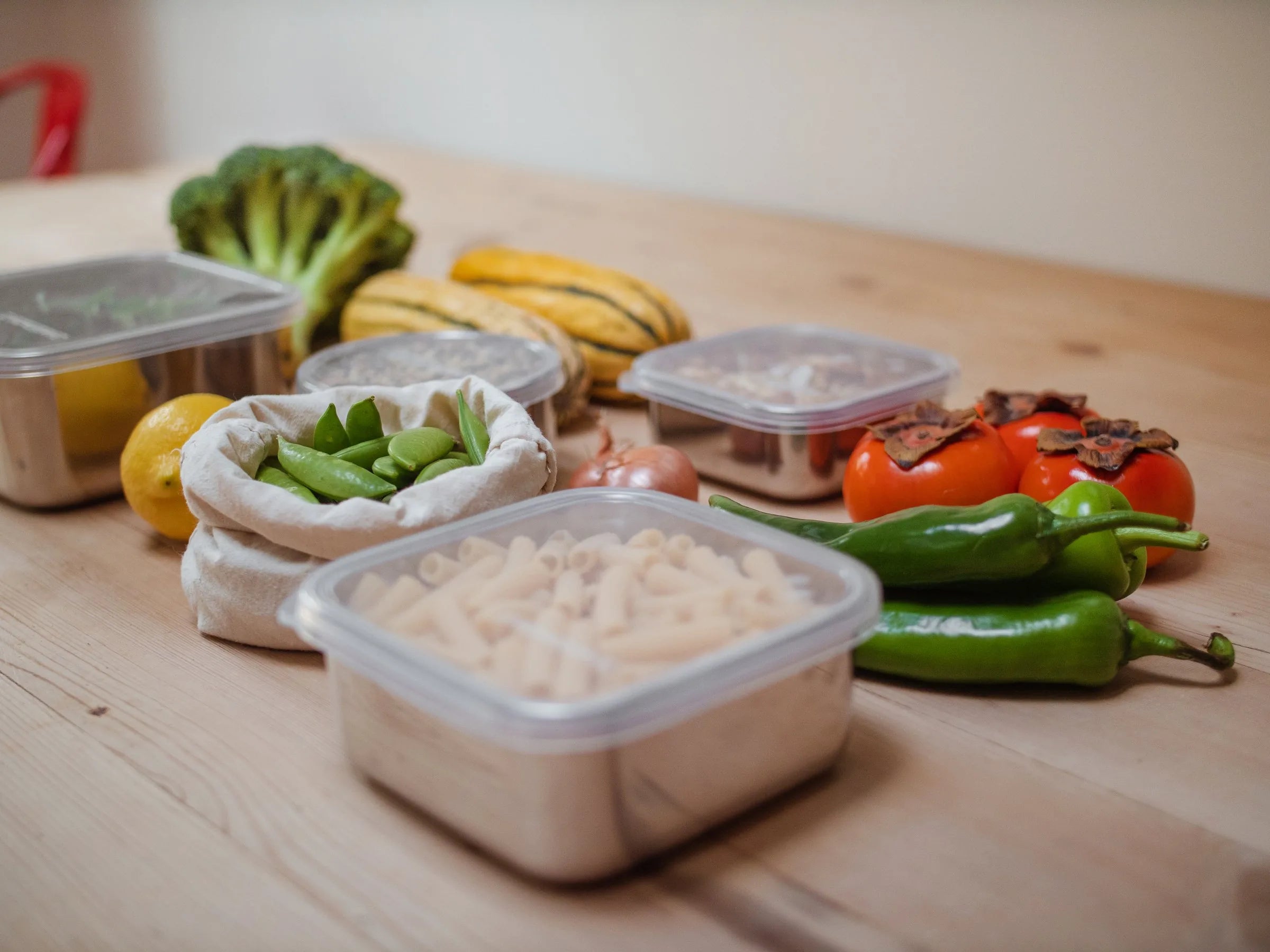 Stainless steel containers with clear lids of pasta and greens on a timber table with fresh vegetables